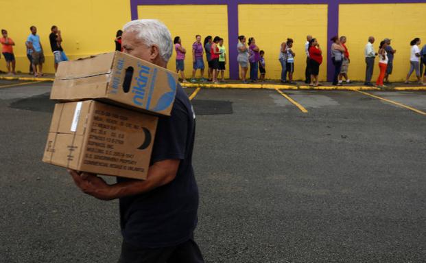 Un hombre camina con dos cajas de alimentos frente a una fila de personas damnificadas por el paso del huracán María. /Thais Llorca (Efe)